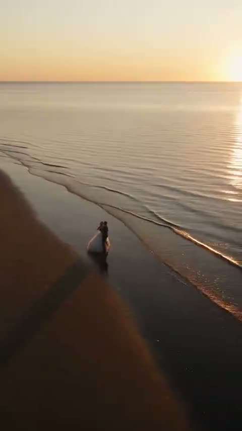 Sunset Serenade: ett brudpar vid vattenbrynet på en lång sandstrand i solnedgången, filmat ovanifrån